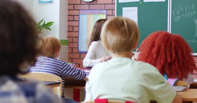 Back view from backs of teens pupils sitting at deskd in classroom and rising hands. Young female Caucasian teacher having class for small kids at school. Education concept.