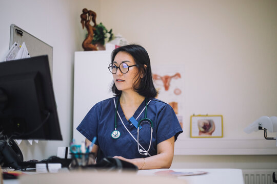 Mature female doctor wearing eyeglasses using computer sitting at desk in hospital