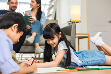 Happy Asian family relaxing on sofa while kid drawing on floor. Little boy girl having fun, friendship between siblings, family leisure time in living room at home.