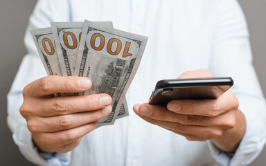 Business and finance concept. Close-up of man holding one hundred dollar bills and using financial mobile application on smartphone while standing indoors on gray background. Selective focus on money