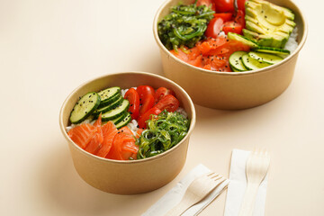 Trendy wholesome dish poke or buddha bowl - rice, wakame seaweed, tomatoes, cucumber, and red fish salmon - in a recycled round carton with sustainable fork, takeaway food concept on beige background