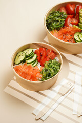 Trendy wholesome dish poke or buddha bowl - rice, wakame seaweed, tomatoes, cucumber, and red fish salmon - in a recycled round carton with sustainable fork, takeaway food concept on beige background