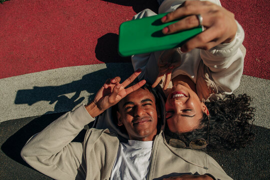 Friends making peace sign while taking selfie through smart phone lying in playground