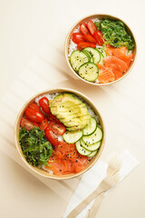 Trendy wholesome dish poke or buddha bowl - rice, wakame seaweed, tomatoes, cucumber, and red fish salmon - in a recycled round carton with sustainable fork, takeaway food concept on beige background