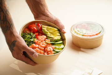 Male hands holding Trendy wholesome dish poke or buddha bowl - rice, wakame seaweed, tomatoes, cucumber, shrimps, avocado - in a recycled round carton with sustainable fork, takeaway food concept