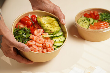 Male hands holding Trendy wholesome dish poke or buddha bowl - rice, wakame seaweed, tomatoes, cucumber, shrimps, avocado - in a recycled round carton with sustainable fork, takeaway food concept