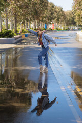 A woman in a denim outfit dances on a wet park alley reflected in a puddle. Spring joyful mood. Walk after the rain