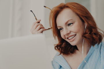 Obraz premium Close up shot of redhead businesswoman makes video call to business partner, has discussion with corporate partner, holds optical glasses, smiles pleasantly, uses earphones. Remote job interview