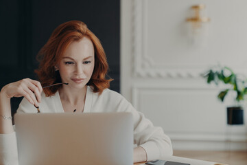 Photo of serious redhead woman journalist concentrated at laptop computer, searches information for...