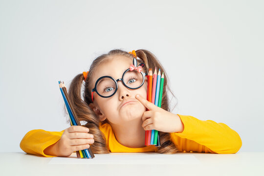 Beautiful cute little preschooler girl with glasses holding colorful pencils and making gesture while looking at camera. Playful child with pencils. Imagination and creativity at school concept. 