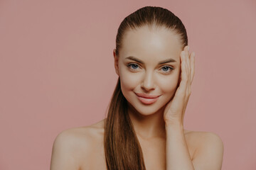 Isolated shot of pretty woman with combed pony tail, enjoys freshness of skin, looks with charming...