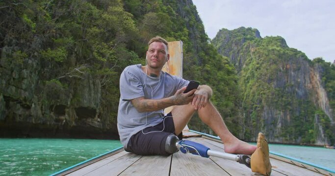 Man with artificial bionic leg prosthesis sitting on the front of long tail boat listening music while enjoying amazing beautiful seascape during summer vacation travel in Phi Phi Islands, Thailand.
