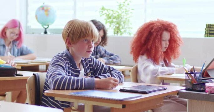 Caucasian clever teen schoolboy sitting at desk in classroom and writing in copybook. Study at schoo conceptl. Smart boy learning at lesson, passing the test and is concentrated.