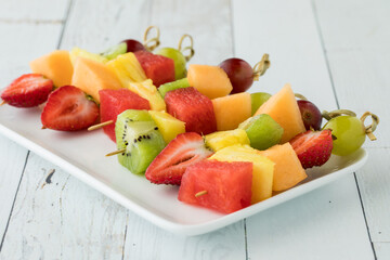 Fresh fruit kebabs on a small platter on a white wash wooden table.