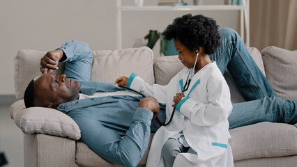 Obraz premium Little cute kid girl plays pretending be doctor adult father lies on couch complains pain symptom playful daughter listening dad with stethoscope child scolds man for bad habits strictly shakes finger