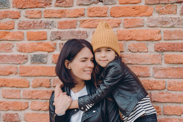 Fototapeta na wymiar Smiling brunette woman and her little daughter embrace with love, have charming smiles, wears black leather jackets, pose against brick wall. Young brunette mother spends free time with female kid