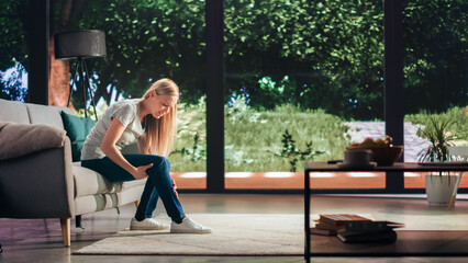 Young Adult Female Had to Sit Down on a Couch at Home Due to Discomfort in a Result of Leg Trauma or Arthritis. Joint and Knee Injury Concept. Massaging the Muscles to Ease the Pain.