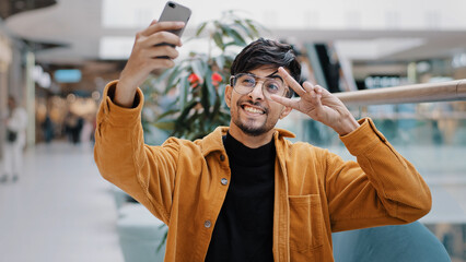 Young happy indian guy blogger holds smartphone recording vlog filming video for blog in mall joyful man waving greeting in webcam on phone video conference online takes selfie photo on mobile camera