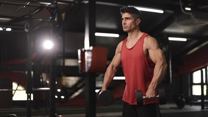 Tough man standing in the gym and pumping his hands with dumbells
