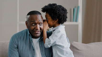 Little cute daughter whispering in dad's ear telling secret mystery says funny joke loving father with kid girl having fun sitting on couch in room burst into laughter laughing joyful man surprised - obrazy, fototapety, plakaty