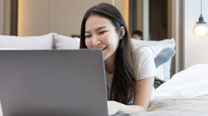 Asian woman uses laptop on bed in modern bedroom at home after she wakes up in the morning,...