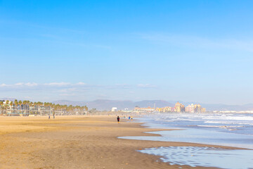 Summer vibes on the sunny autumn beach of Malvarrosa in Valencia, Spain. Vast expanses of smooth fine sand on the sea coast attract vacationers to solitary walks along the bubbling foamy waves.