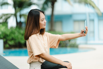 Fototapeta na wymiar A close-up shot of a young Asian tourist taking a selfie with her cell phone happily at the poolside of her hotel on a weekend.