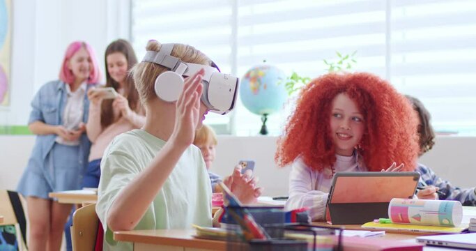 Little Caucasian boy pupil in VR headset sitting at desk in classroom and having virtual reality learning experience. Interactive study concept. Schoolboy in VR glasses moving hands.