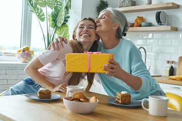 Happy senior woman receiving a gift box from her adult daughter while both sitting at the kitchen