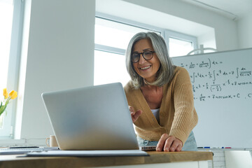 Mature woman teaching mathematics while standing near the whiteboard and looking at laptop - obrazy, fototapety, plakaty