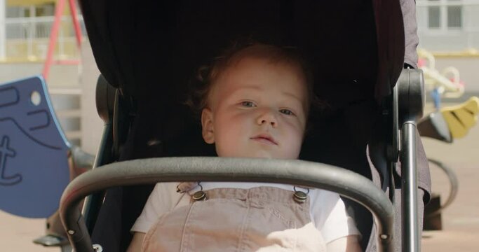 Baby girl sits in black carriage on children playground. Little daughter in pram looks around. Baby on walk with family on summer day closeup