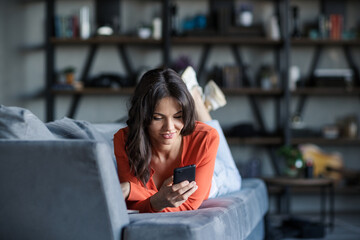 Smiling attractive brunette woman lying on sofa at home and typing sms on cell phone. Beautiful female exchanging text messages on mobile phone