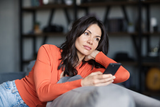 Attractive brunette woman sitting on sofa at home and thinking while text messaging on cell phone. Beautiful female contemplating over sms on mobile phone