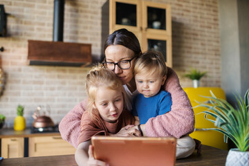 Happy young mother and two kids, little daughter and baby son, watching cartoons on tablet computer. Loving family enjoying time together at home, mom hugging children