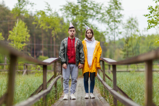 Unhappy Disappointed Young Couple In Party Hats Standing On Wooden Path In Countryside And Looking Sadly At Camera In Rain. The Party Is Over, Celebration Failed, Hike Gone Wrong