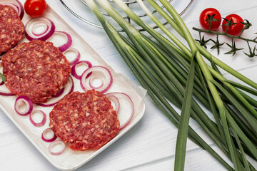 Green onion and tomato sprig. Raw beef burgers with onions on a plate.