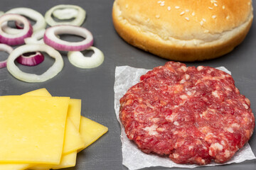 Sliced cheese, raw burger on paper, sesame bun and chopped onion on table.