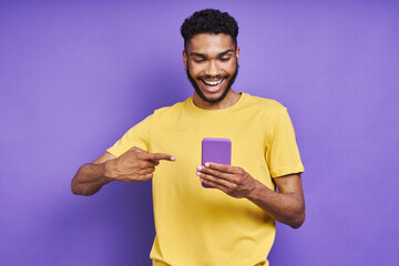 Cheerful African man pointing his smart phone while standing against purple background