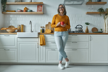 Cheerful senior woman holding cup and smiling while standing at the domestic kitchen