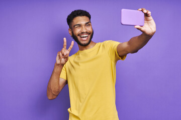 Playful African man making selfie by smart phone and gesturing against purple background
