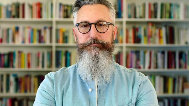 University lecturer and literature professor standing in front of a library bookshelf. Portrait of a serious and intelligent intellectual professor, researcher or author with a grey beard and glasses