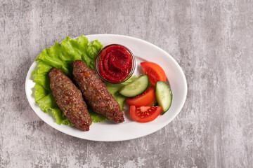 The national dish lyulya-kebab with vegetables and lettuce leaves with red sauce in a plate on a gray background. Horizontal orientation, top view, no face, copy space