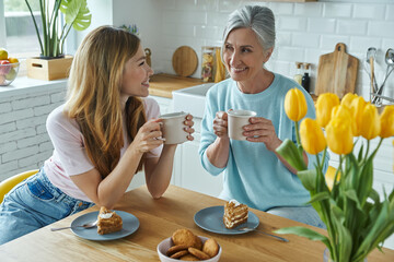 Senior mother and her adult daughter enjoying hot drinks and sweet food at the kitchen
