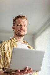 Portrait of bearded middle aged man holding open laptop computer and looking at camera standing indoors. Male working online from home or browsing Internet on pc