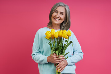 Happy senior woman holding bunch of tulips while standing against pink background