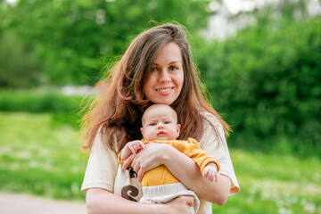 A young mother gently holds a newborn baby dressed in a yellow suit in a green summer park. Kid in mother's arms on summer day in sun in park.