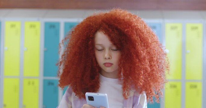 Portrait of cute Caucasian small girl with red curly hair standing at school corridor among people on a break, using smartphone and looking at camera. Little teen schoolgirl with a gadget.