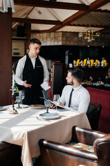 A young businessman in a fine restaurant examines the menu and makes an order to a young waiter in a stylish apron. Customer service. Table service in the restaurant.