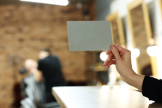 female hand holding grey postcard or bossiness card with mock up on stylish barbershop or beauty salon background. blurred barber man is cutting and shaving the beard o client.