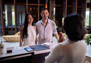 Young couple walking in lobby and arriving at hotel reception while front desk employee welcoming and greeting them - 517535797